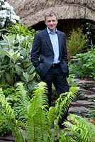 Portrait of Chris Beardshaw in Furzey Garden. Fern in foreground is Matteucia struthiopteris. Other plants include Rhododendron 'Hoppy', Geranium palmatum, Rhododendron 'Cunninghams White', and Primula 'Apple Blossom'.