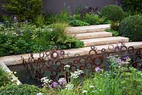 A garden inspired by the Arts And Crafts Movement. Formal paths and terraces combine with a water channel. Focal point is an energy wave sculpture made from copper rings. Planting style is woodland edge with buxus domes - The M & G Garden