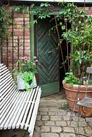 Granite patio with open wooden door, white bench, containers and a bag with Cleome 'Senorita Rosalita', Cymbopogon citratus 'Tasty Lemon' and Glechome hederacea 'Variegata' 