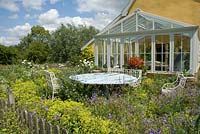 Conservatory on west side of Wood Farm, paved area with Alchemilla mollis and Geraniums. Red Pelargonium in a pot with garden furniture. June