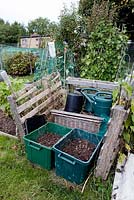 Neat, rustic fenced in storage area on allotment with compost in green recycling bins and plastic watering cans behind - Fortis Green Allotments, London Borough of Haringey 1