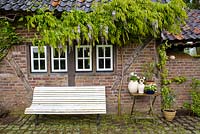 White painted wooden bench on a small patio by the summer house with Wisteria growing up a brick wall, De Carishof