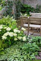 Wooden benches on stone patio with brick wall and water trough and fountain and attractive green and white planting with Ajuga, Hosta, Geranium, Fatsia, Pulmonaria, Hydrangea, Hedera and ferns in the 'Enchantment' garden