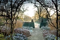 Formal garden in winter with borders of Sedum, Calamagrostis acutiflora and Buxus viewed through arch of climbing Rosa - Winfield House