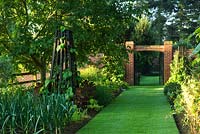 Wildlife conservation garden with long mown grass path, obelisk, brick arch and metal gates