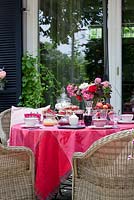 On a terrace, wicker chairs next to a red linen clothed table that is prepared for breakfast with fruit, jam and a flower bouquet