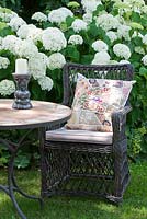 Wicker armchair with embroidered patchwork cushion showing a butterfly next to mosaic bistro table with candleholder and a white Hydrangea arborescens 'Annabelle' in the background