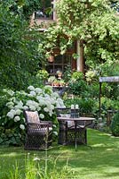 Two wicker armchairs with embroidered patchwork cushions and a bistro table with candle holders against a backdrop of white flowering Hydrangea arborescens 'Annabell'.

