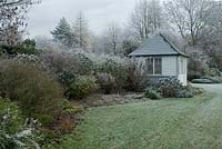 Shrub and perennial border including Euphorbia, Roses and Hebes with a wooden painted summer house. Frost in December