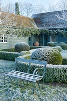 View to house with wrought iron garden seat. Yew and box topiary and dwarf hedges on frosty morning in December - The Mill House, Little Sampford, Essex
