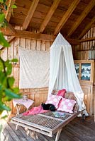 Under a wooden porch, a black cat is sitting on a sunbed with cushions and a mosquito net