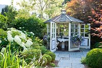 White wooden garden pavilion at end of flagstone path. Plants are Buxus, Hosta, Hydrangea arborescens 'Annabell', Petunia Surfinia and Prunus laurocerasus