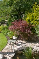 Seating area on limestone patio by pond with mature trees and shrubs in borders beyond. The garden is open for the National Garden Scheme in Dalton Hamlet, Cumbria