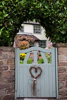 Pale blue cottage garden gate with heart decoration, set into stone wall with Prunus laurocerasus 'Rotundifolia' hedge - Garden Neighbours