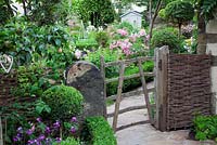 Small cottage garden seen through rustic wooden gate with old stone pillar and woven willow fence. Buxus circle with decorative metal obelisk. Plants include Rosa 'Ballerina', Ligustrum standard trees, low Buxus hedge and Apple tree - Garden Neighbours
