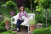 Liz, cottage owner sits with Archie on painted bench on brick terrace with Buxus balls and low Buxus hedge, standard Bay trees and potted grasses - Garden Neighbours