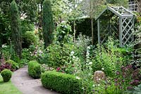 Small cottage garden with stone pathway edged by Buxus low hedging. Two sentinel Juniper  'Skyrocket', wooden bower seat, Astrantia 'Claret', Rosa 'Tuscany Superb', Campanula glomerata and Campanula persicifolia. Garden Neighbours