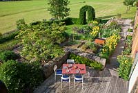 A varying rural garden with garden furniture on a wooden terrace, a stone wall, a clipped hornbeam arch and edged vegetable patches. Plants include Buxus, Fagus sylvatica and Iris germanica
