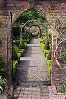Brick wall and entrance with iron gate, long gravel path,borders framed with box hedging and box cones, with arbour. 
