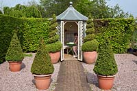 Summerhouse with twisted and pyramid topiary box in terracotta pots on gravel and brick pathway 