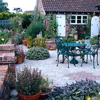 Patio of crazy paving and bricks with metal table and chairs and container planting throughout