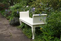 Decorative white wooden bench in spring garden with Symphytum grandiflorum, Paeonia lactiflora and Camassia cusickii 