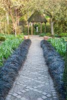 A path of stone setts and gravel in the Canal Garden is framed by black Ophiopogon planiscapus 'Nigrescens' and leads towards the Dell Garden with white drinking fountain as focal point