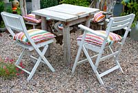 Seating area with small square table made of drift wood and cushioned chairs on gravel in the corner of cottage garden