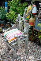 Chair made of driftwood on gravel surface in corner of cottage garden surrounded by vintage collection of galvanised planted containers and fishing floats against wooden fence