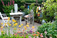 Seating area with small square table and chairs made of drift wood on graveled corner of cottage garden with collection of vintage galvanised containers fishing floats and planting including Lupinus Primula and Geranium 