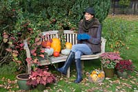 Woman sat on garden bench with autumnal display of pumpkins. 'Crown Prince', 'Mammoth', 'Uchiki Kuri' and basket of Gourds.