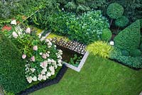 View to the garden from top level of the house. Small pond with granite setts border  amongst Buxus,   Rhododendron 'Cunningham's White', Carex oshimensis 'Everillo', Pachysandra terminalis, Lysimachia nummularia 'Aurea', Hydrangea paniculata 'Limelight', Ilex crenata 'Golden Gem', Ophipogon planiscapus 'NIgrescens'.