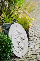 Engraved concrete plaques with angels figures leaning against flower pots with grasses on granite setts path.