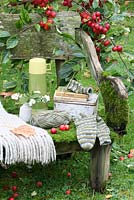 A rustic, moss covered bench with thermos flask, lunchbox, woollen throw, sock knitting, vase of snowberries and Observer books