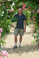 Phil Mizen with a bucket of herbs:  Artemisia dracunculus - French tarragon, Anethum graveolens - Dill and Rumex acetosa - Sorrel under the rose arch with Rosa 'New Dawn' and Rosa 'American Pillar' at Langham Herbs, Walled Garden, Suffolk. July