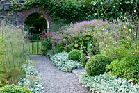 Gravel path leading to a gate and archway in a wall and borders planted with Geranium psilostemon, Stachys byzantina, Aquilegia Foeniculum vulgare and clipped Buxus sempervivum balls 