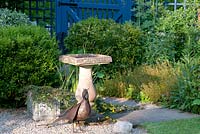 Stone bird bath and metal bird ornament in gravelled area with adjacent blue wooden gate and bed with shrubs and perennials 