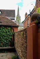 Small town garden with Ivy clad wall, wooden bench and box ball beneath a flint and brick wall. York stone paving and a Church spire in background