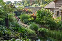 View from wooded slopes that surround the house and garden full of exuberant planting that features tree ferns, cannas, ricinus and hostas with walled garden beyond. 