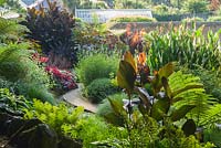 View from wooded slopes that surround the house and garden full of exuberant planting that features tree ferns, cannas, ricinus and hostas with walled garden beyond.