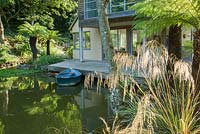 New build house at the end of the lake surrounded by grasses and tree ferns, with rowing boat moored beside the deck. 