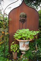 Metal feature in the Parterre Garden with cherub's head and urn with pelargonium. Whimble Garden and Nursery, Kinnerton, Powys, Wales