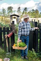 Man standing at the entrance gate of the garden holding trug of harvested vegetables.