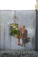 Planting display in vertical container hanging on wall with painting of children