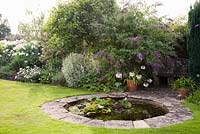 Decorative stone circular pond in summer with fountain and planting including Phlox, Phalaris arundinacea, Buddleia and Agapanthus