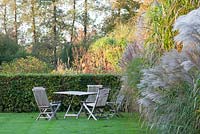 Wooden furniture on lawn next to mixed grass border - Miscanthus sinensis 'Herman Muessel', Miscanthus sinensis 'Professor Richard Hansen'