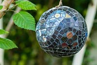 Mosaic ball, designed by Anne Cardwell, hanging from a Betula - silver birch tree in town garden