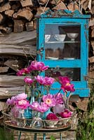 Backed by a wood pile and a blue cupboard with tea cups, arrangement of pink and red peonies on an antique metal desk