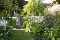 Central stone statue on grass pathway with Hesperis, Aquilegia and Buxus balls either side and pergola and greenhouse behind. The Old Rectory, Dorset