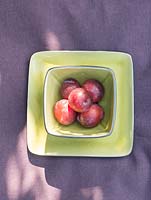Display of green tableware with red plums in the bowl on purple tablecloth.
